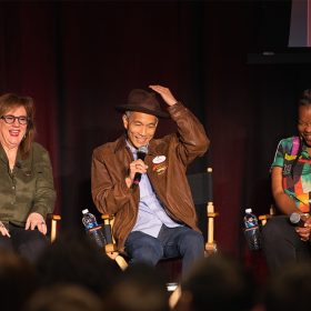 Three panelists sit on stage in director's chairs during the first D23 Spotlight Series on February 22 highlighting the Indiana Jones Adventure attraction. The individual in the center wears a brown leather jacket, a fedora, and holds a microphone, evoking the style of Indiana Jones. The panelist on the left wears an olive green shirt, while the one on the right sports a colorful patterned top.