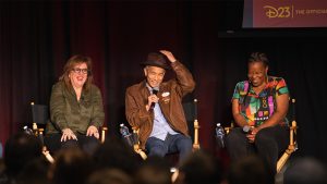 Three panelists sit on stage in director's chairs during the first D23 Spotlight Series on February 22 highlighting the Indiana Jones Adventure attraction. The individual in the center wears a brown leather jacket, a fedora, and holds a microphone, evoking the style of Indiana Jones. The panelist on the left wears an olive green shirt, while the one on the right sports a colorful patterned top.