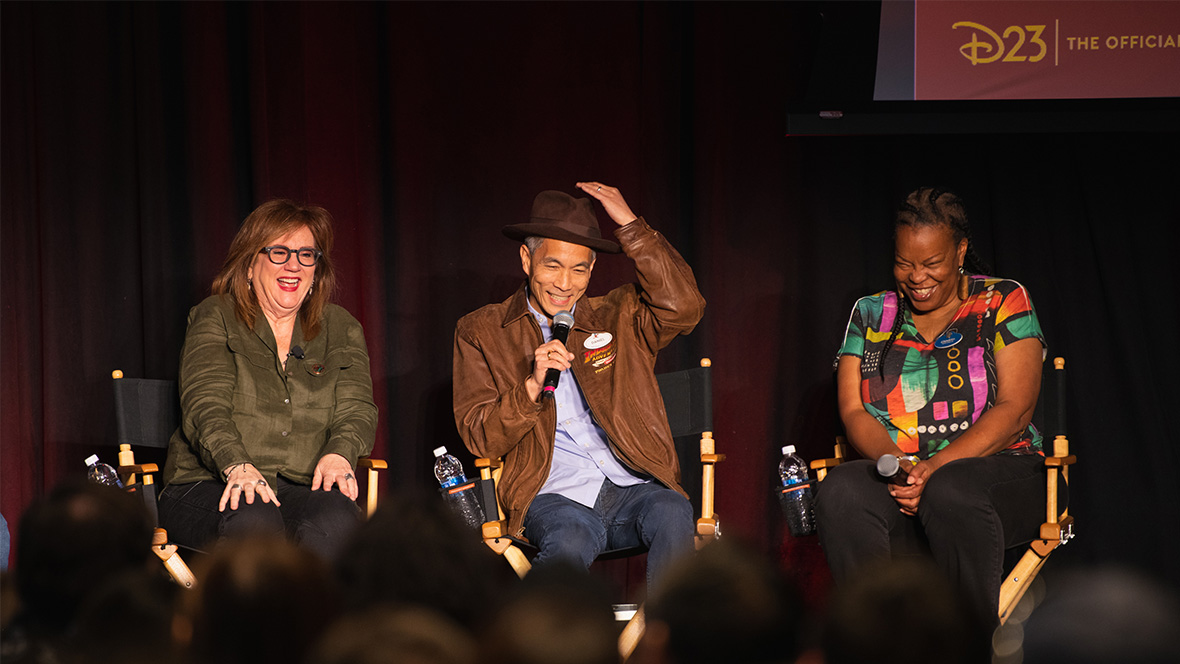 Three panelists sit on stage in director's chairs during the first D23 Spotlight Series on February 22 highlighting the Indiana Jones Adventure attraction. The individual in the center wears a brown leather jacket, a fedora, and holds a microphone, evoking the style of Indiana Jones. The panelist on the left wears an olive green shirt, while the one on the right sports a colorful patterned top.