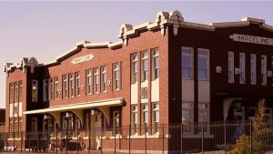 A historic brick building with white trim labeled 'Marceline' on both wings, representing the former train depot in Marceline, Missouri. The two-story structure features large rectangular windows and arched entrances, with a vintage architectural style. A black iron fence surrounds the property, and a clear sky provides a warm backdrop. This site is significant for its connection to Walt Disney’s childhood and the inspiration behind Main Street, U.S.A.