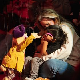 Image from D23 Day at Bob Baker Marionette Theater featuring two adult guests—with a child seated in the lap of one of the guests—on red flooring, interacting with a rabbit marionette. The rabbit marionette's ears are down under a purple beret, and it’s wearing a yellow coat with its arms raised towards them. In the foreground are other guests looking at the interaction.