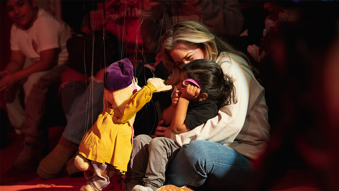 Image from D23 Day at Bob Baker Marionette Theater featuring two adult guests—with a child seated in the lap of one of the guests—on red flooring, interacting with a rabbit marionette. The rabbit marionette's ears are down under a purple beret, and it’s wearing a yellow coat with its arms raised towards them. In the foreground are other guests looking at the interaction.