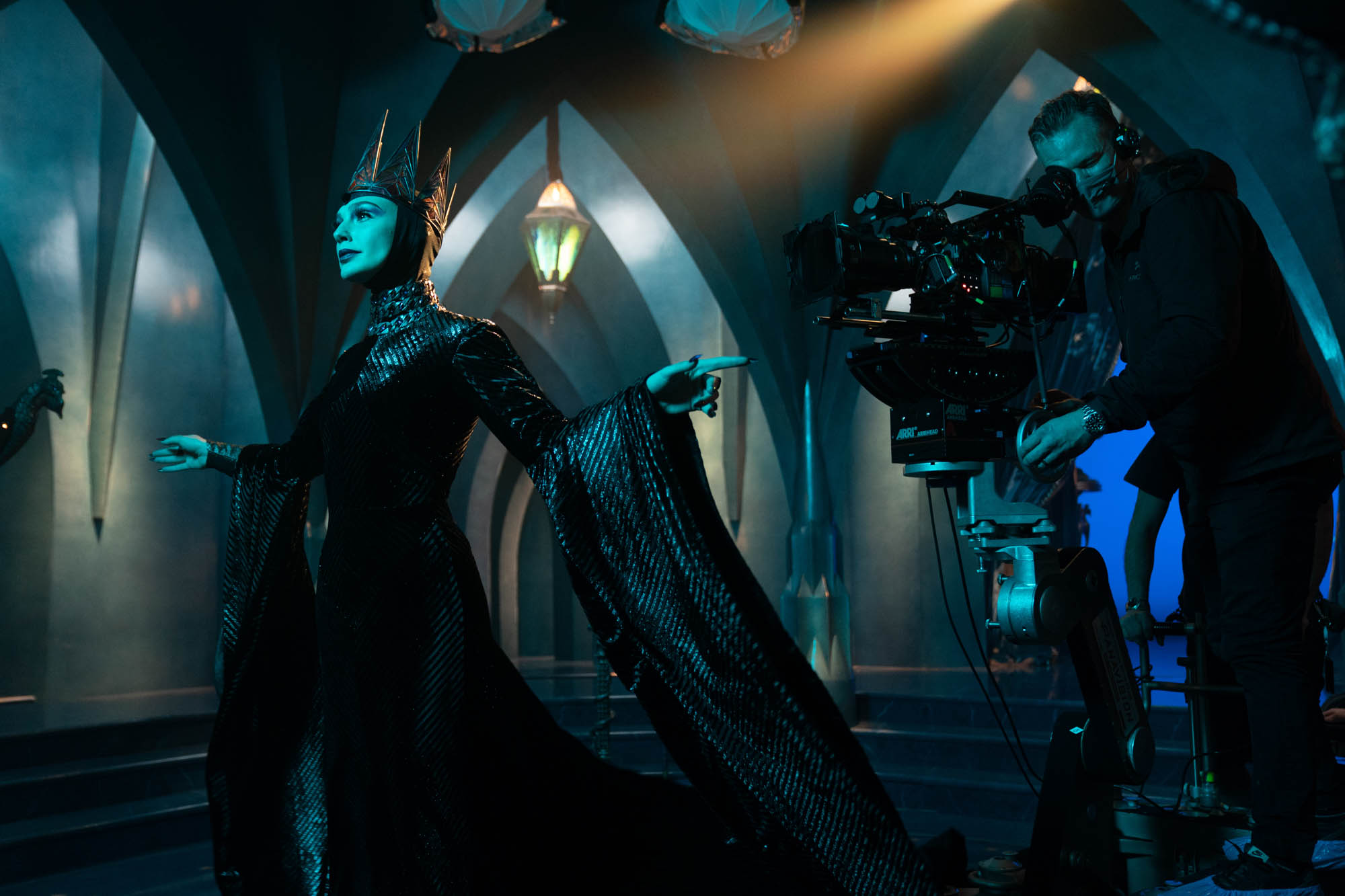 Gal Gadot as the Evil Queen wears a dramatic high-necked glittering dark long-sleeve gown and a steely crown atop her head. She glances at an unseen mirror to the left. A cameraman is positioned at the right, capturing the scene.