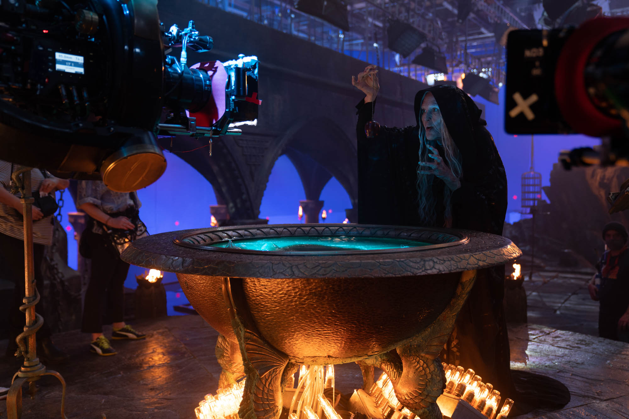 Gal Gadot as the old hag stands in a hooded dark cloak before her glowing cauldron as she prepares the poison apple. Cameras are positioned at both sides of the image, recording the scene.