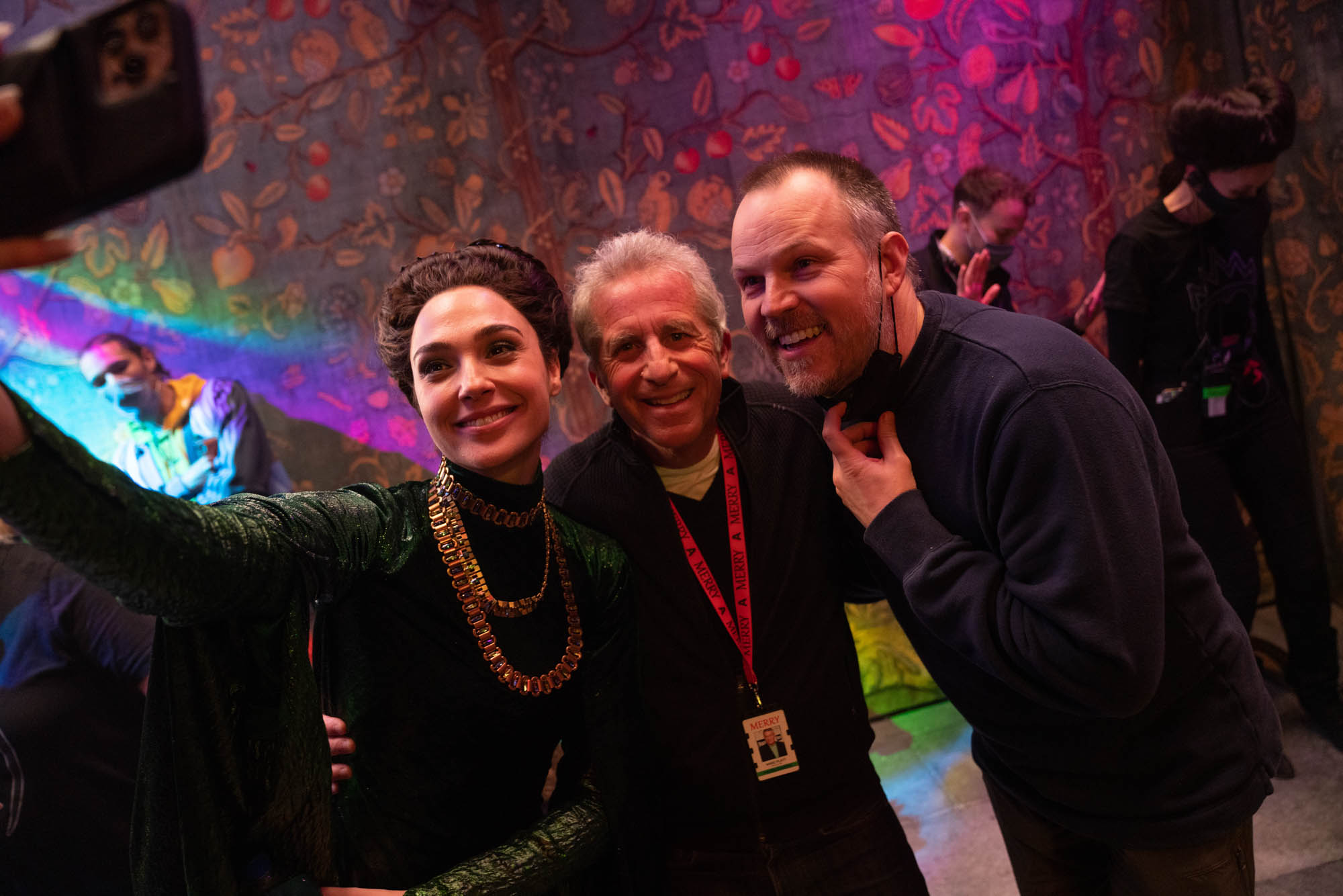 Gal Gadot is seen on set taking a selfie with producer Marc Platt at center and director Marc Webb at left. She wears an emerald green dress with a multiple strand chunky gold and silver chain necklace. Everyone smiles in the direction of the phone’s camera lens.