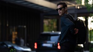 Rami Malek appears in a scene from The Amateur as Charlie Heller. He’s standing with his left side facing the camera, in the process of putting on a backpack. He’s turned to his left, so we can see his face, and he looks apprehensive. He’s wearing dark sunglasses and all black clothes. Beyond him, out of focus, is what appears to be the entrance to a hotel or swanky apartment complex.