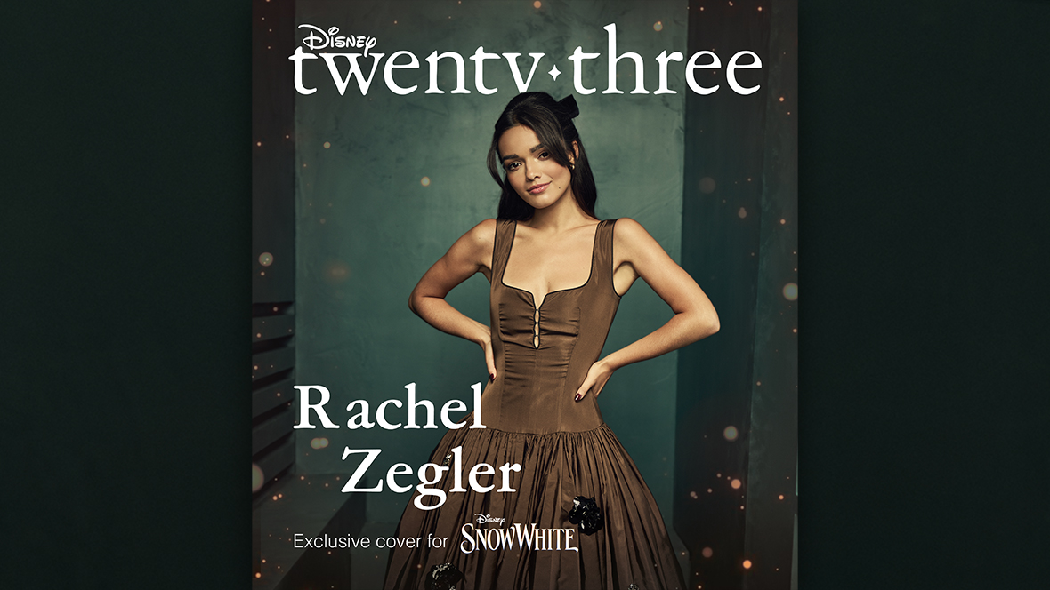 Rachel Zegler stands with manicured hands in a deep plum at her hips with a slight smile on her face as she looks at the camera. Her dark hair is half pulled back with a large black bow partially visible. She wears a sleeveless, bodice-style rust-colored dress with one glossy black rose below the hip at right. The background is a teal green with specks of gold scattered throughout. The copy reads: “Disney twenty-three” at top. With “Rachel Zegler Exclusive cover for Disney Snow White” at lower left.