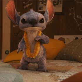 Stitch from the 2025 live-action version of Lilo & Stitch. He is wearing a yellow lei and standing on a bed with green and yellow pillows and a pink stuffed poodle. Behind him is a wall with Polaroid pictures and drawings of the constellations.