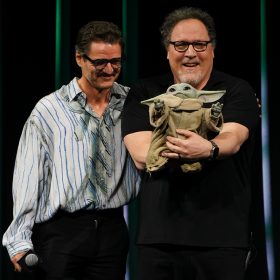 On stage at Star Wars Celebration Japan, Pedro Pascal looks on as Disney Legend Jon Favreau holds Grogu.