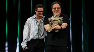 On stage at Star Wars Celebration Japan, Pedro Pascal looks on as Disney Legend Jon Favreau holds Grogu.