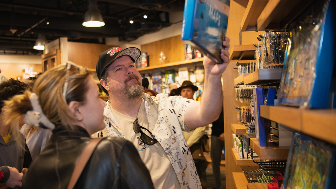 D23 Members browse merchandise at an exclusive Disney shopping event. A man in a printed shirt and baseball cap examines a book or collectible item from a shelf while a woman beside him, wearing a leather jacket and Minnie Mouse ear scrunchie, looks on. The store is filled with themed products and display shelves, with other shoppers visible in the background enjoying the event.
