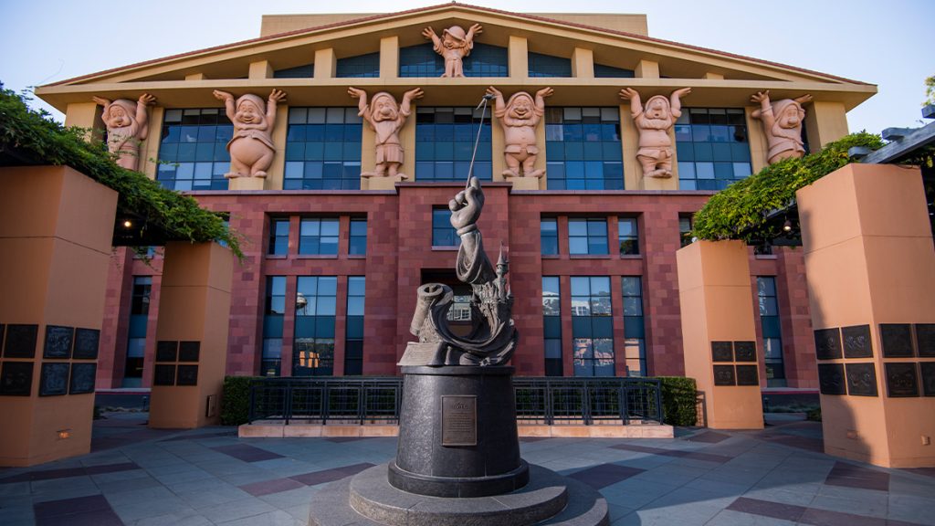 Front view of the Team Disney Building at the Walt Disney Studios in Burbank, California. The upper facade features massive sculpted figures of the Seven Dwarfs holding up the roof, a tribute to Snow White and the Seven Dwarfs. In the foreground is the Disney Legends Plaza with a statue of Mickey Mouse's gloved hand holding a wand above a swirl of magical elements, including Cinderella&rsquo;s Castle and film reels. The surrounding pillars display plaques honoring Disney Legends.