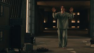 Bob from Thunderbolts* (2025) standing in a gray room with lights behind him and papers and boxes strewn over the floor. He is wearing a light bluish-gray inmate uniform and holding his hands up in a halting gesture.