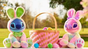 (L-R) Stitch and Angel plushes sitting on a white bench with a grassy field in the backdrop. The plushes are dressed in tie-dye bunny costumes with a basket filled with Easter eggs.