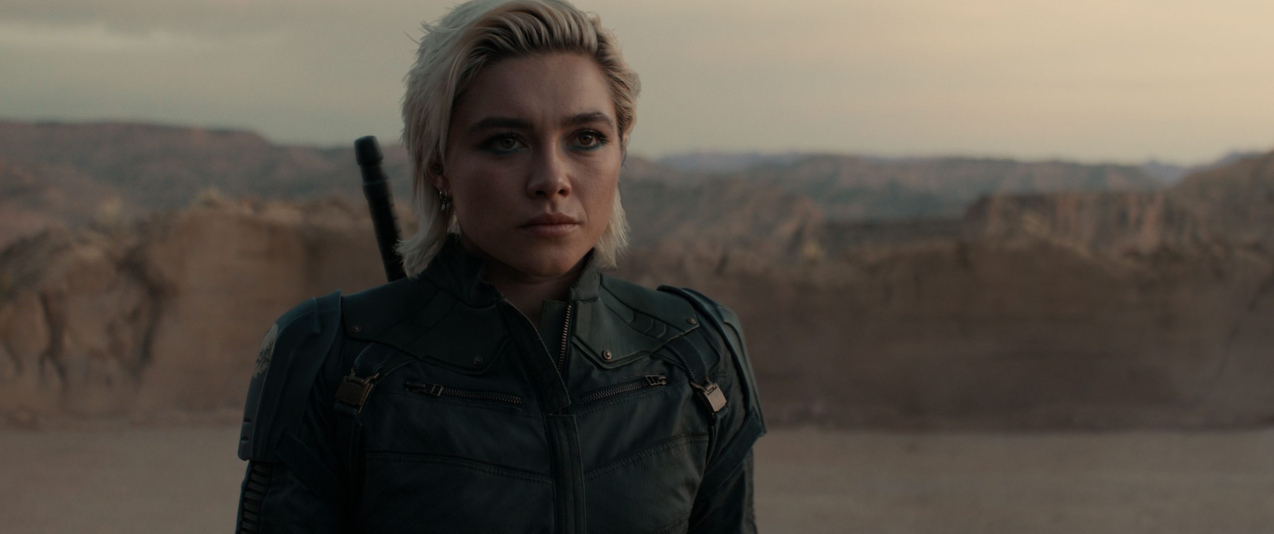 In an image from Marvel’s Thunderbolts* The New Avengers, Yelena Belova (Florence Pugh) is wearing a black Super Hero suit and standing in rocky terrain. It’s dusk, and she’s looking off camera to the right, with a serious expression.
