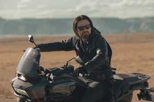In an image from Marvel’s Thunderbolts* The New Avengers, Bucky Barnes (Winter Soldier) is wearing a black leather outfit and sunglasses and is on the back of a motorcycle, surrounded by rocky terrain. It’s daylight and he’s looking at something off camera to the right.
