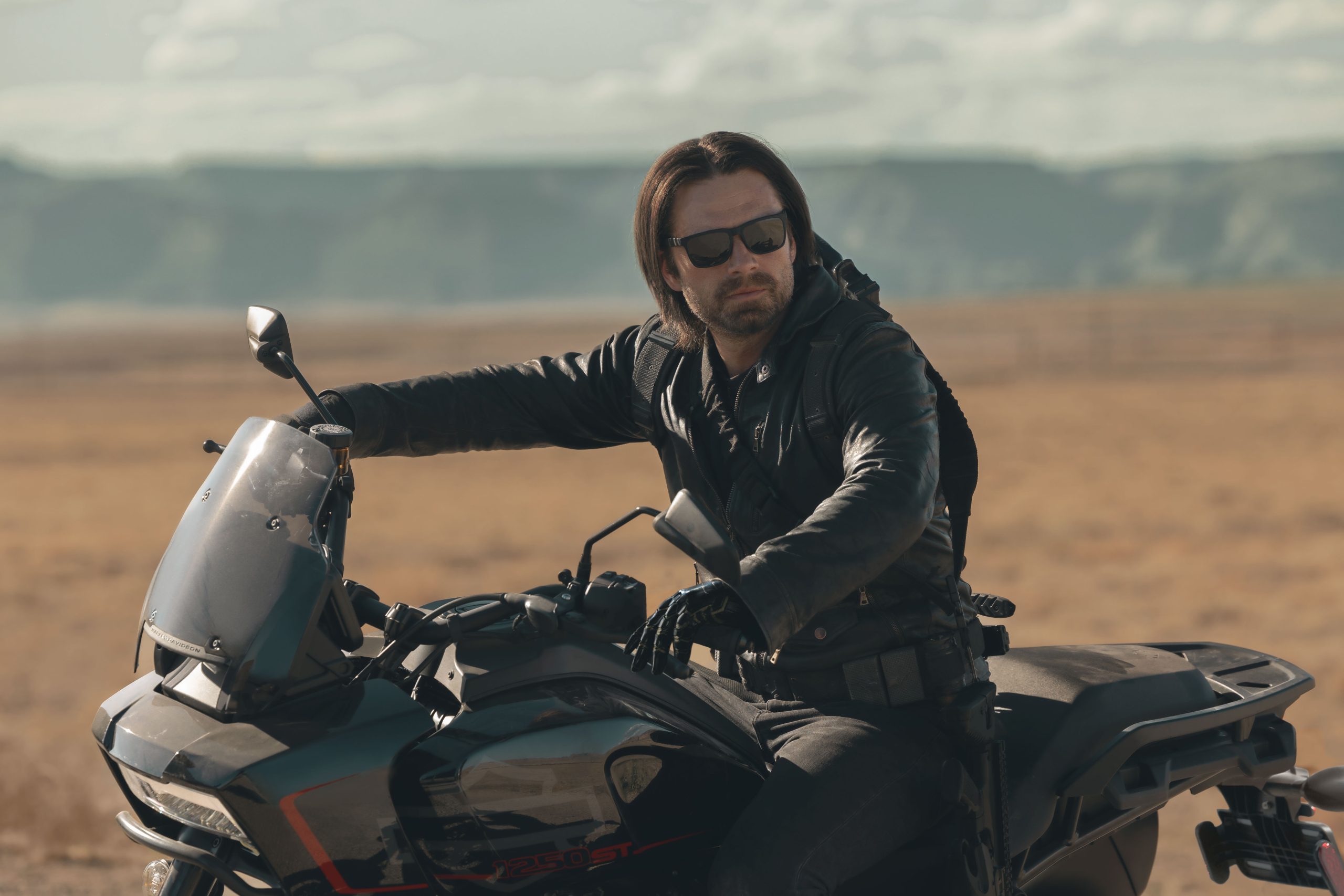 In an image from Marvel’s Thunderbolts* The New Avengers, Bucky Barnes (Winter Soldier) is wearing a black leather outfit and sunglasses and is on the back of a motorcycle, surrounded by rocky terrain. It’s daylight and he’s looking at something off camera to the right.