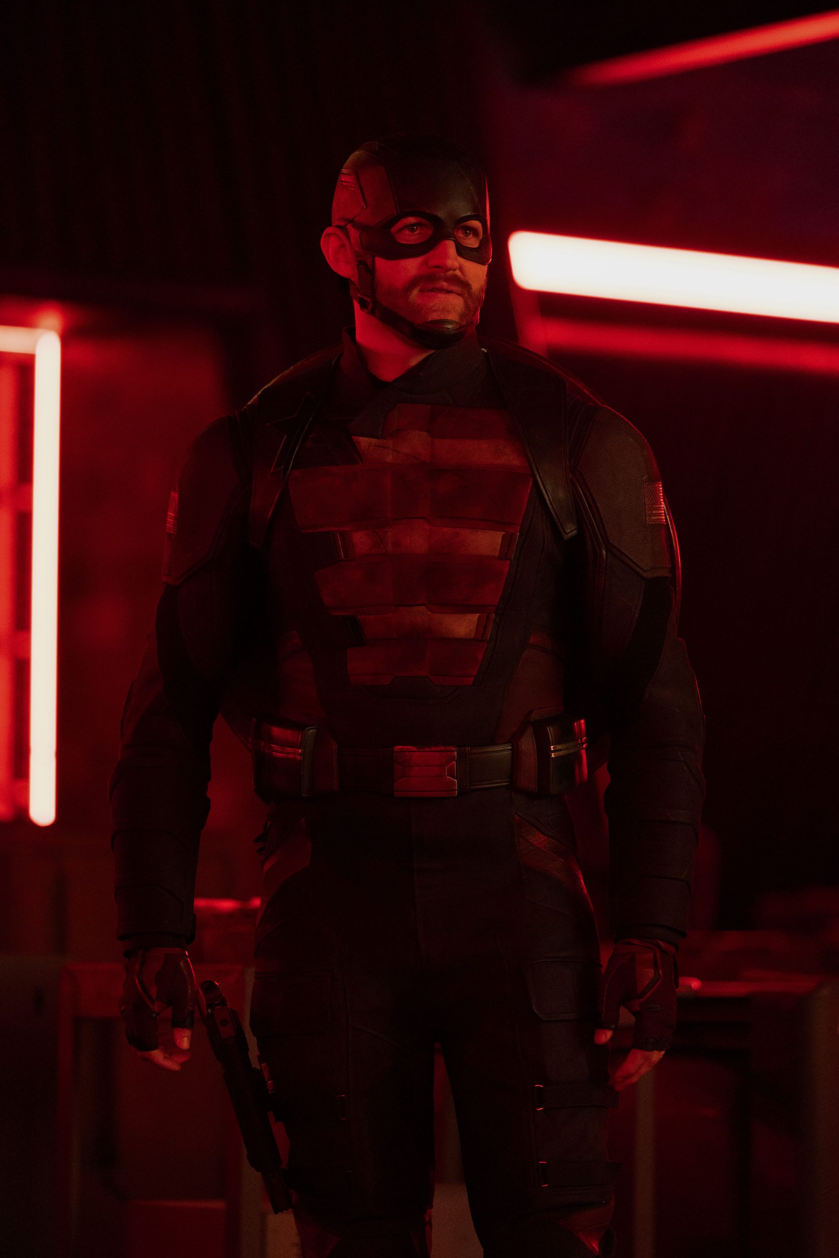 In an image from Marvel’s Thunderbolts* The New Avengers, John Walker (U.S. Agent), in his Super Hero suit, is standing in a darkened hallway somewhat illuminated by long red neon lights. He is looking at something off camera to the right.