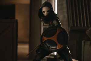 In an image from Marvel’s Thunderbolts* The New Avengers, Antonia Dreykov (Taskmaster), in her Super Hero outfit, is holding a shield and weapon and standing near several boxes, to the left, in a dimly lit room. Her face is covered by a white mask and dark cloak.