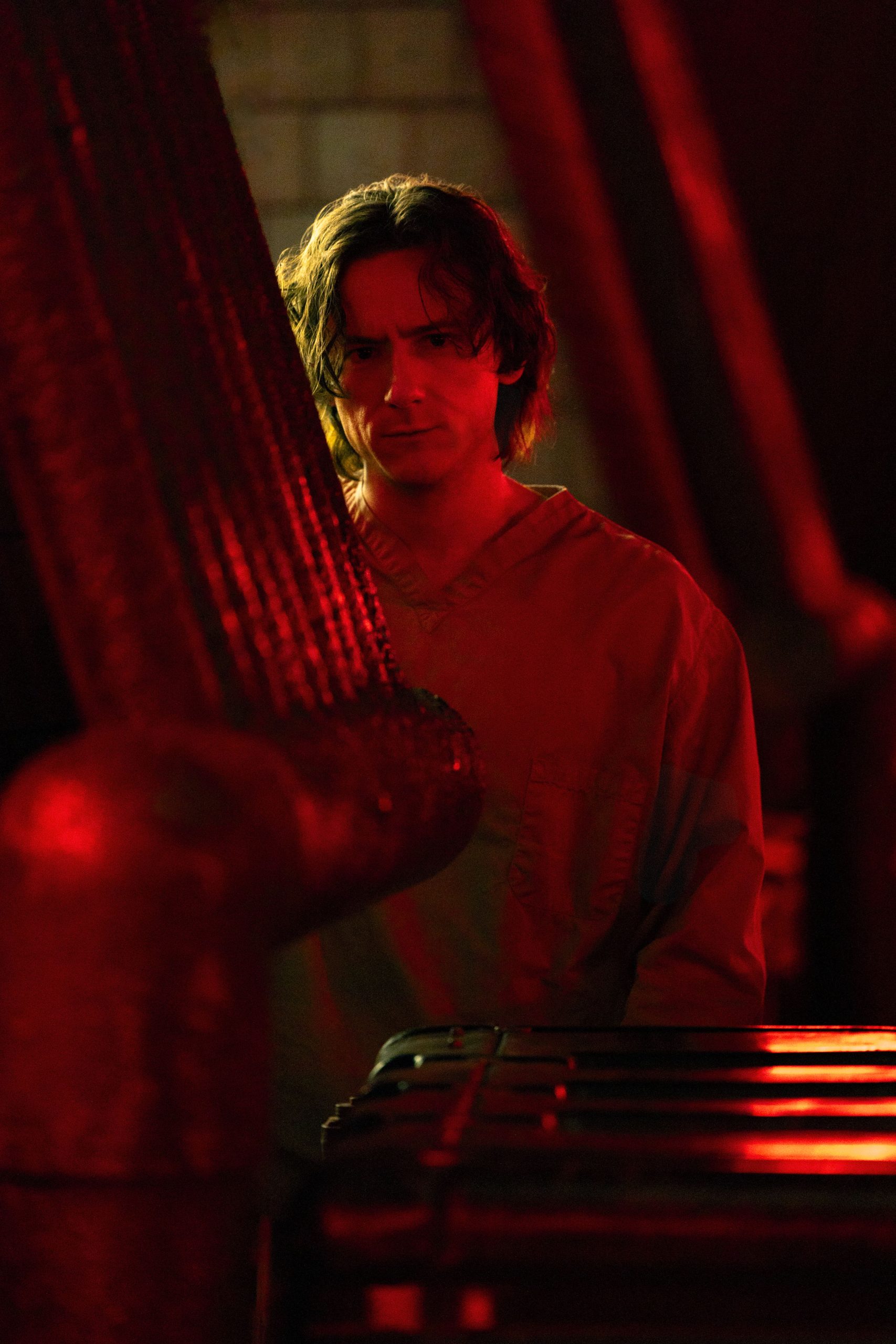 In an image from Marvel’s Thunderbolts* The New Avengers, Bob (Lewis Pullman) is wearing a light bluish-gray inmate uniform and standing behind a column and black plastic boxes, looking at something behind the camera’s vantage point. The room he’s in is dimly lit by red neon lights.