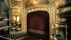 Interior of the historic New Amsterdam Theatre in New York, showcasing its opulent Art Nouveau architecture with ornate balconies, intricate carvings, and a grand proscenium framing deep red stage curtains.