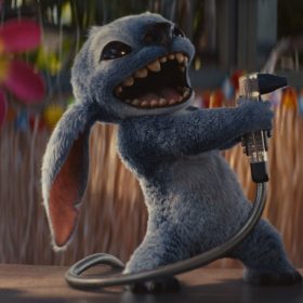 Stitch, a small blue alien with large ears and sharp teeth, energetically holds a soda dispenser hose like a microphone, appearing to sing or shout. He's standing on a counter with a colorful Hawaiian-themed background, including flowers and a straw skirt decoration.