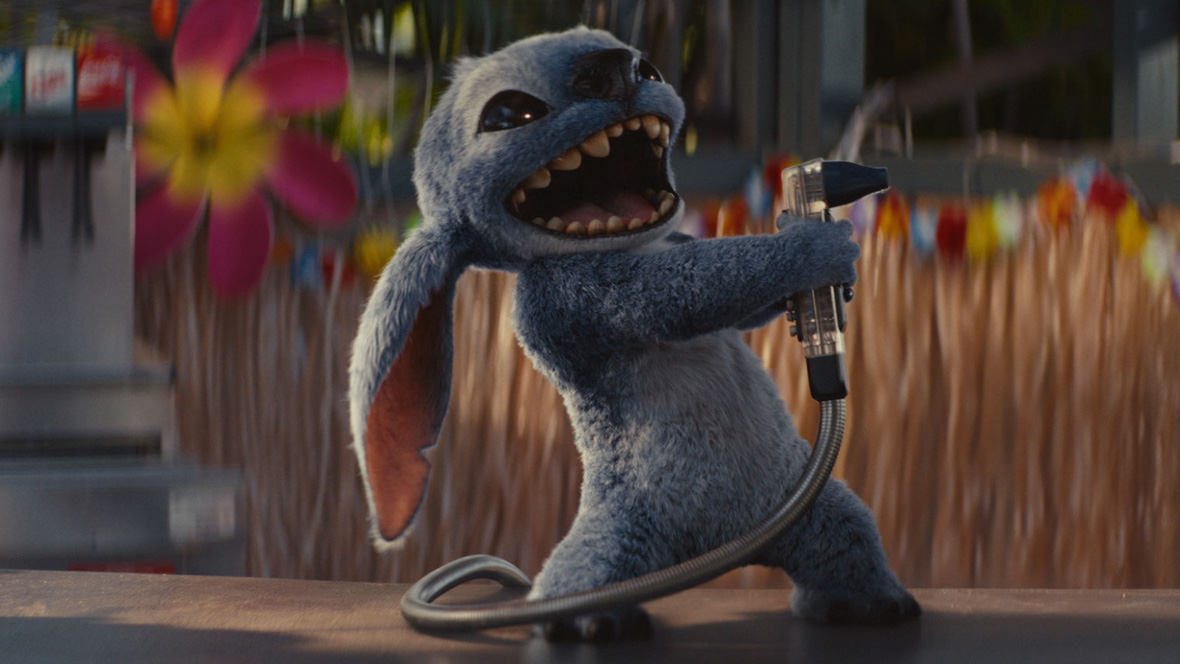 Stitch, a small blue alien with large ears and sharp teeth, energetically holds a soda dispenser hose like a microphone, appearing to sing or shout. He's standing on a counter with a colorful Hawaiian-themed background, including flowers and a straw skirt decoration.