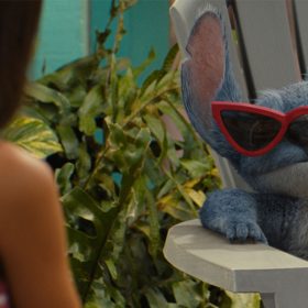 Stitch, a blue alien with large ears and furry texture, lounges confidently in a white outdoor chair wearing red cat-eye sunglasses. Lilo is in a pink floral dress is partially visible in the foreground, with tropical plants and colorful decor in the background.
