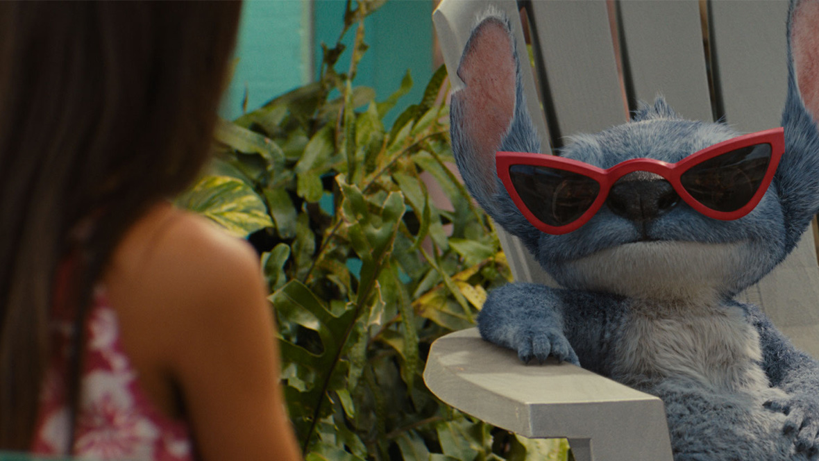 Stitch, a blue alien with large ears and furry texture, lounges confidently in a white outdoor chair wearing red cat-eye sunglasses. Lilo is in a pink floral dress is partially visible in the foreground, with tropical plants and colorful decor in the background.