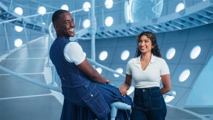 : The Doctor (played by Ncuti Gatwa) and his companion Belinda Chandra (played by Varada Sethu) inside the TARDIS from Doctor Who. The Doctor is wearing a blue and white striped vest with matching pants and smiling at the camera, Belina is wearing a white collared shirt and dark wash jeans and smiling at The Doctor.