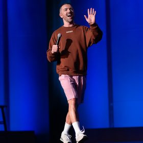 In a still from his new Hulu special, comedian Matteo Lane is onstage in front of a blue background. He is wearing a brown sweatshirt, pink shorts, and white sneakers while waving his left hand and holding a microphone in his right hand.