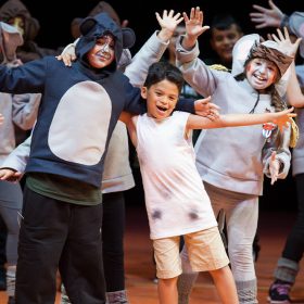 A group of students is posed on stage—all dressed in street clothing (like hoodies, dresses, and T-shirts) made to look like different animals—during a Disney Musicals in Schools performance. Their arms are outstretched and they’re all smiling.