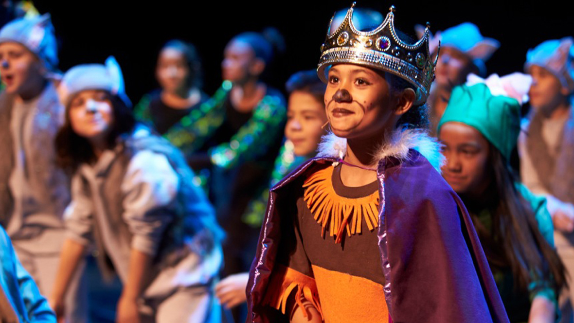 A group of students is posed on stage—all dressed in street clothing (like hoodies, dresses, and t-shirts) made to look like different animals—during a Disney Musicals in Schools performance. One student is featured toward the front; they are wearing a crown and a cape