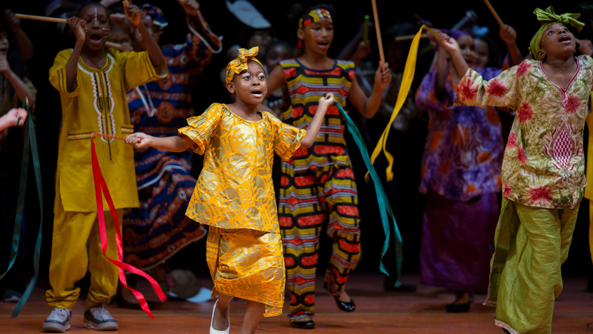 A group of students is posed on stage during a Disney Musicals in Schools performance. They are in the middle of a dance number, and many are holding drumsticks or other sticks with ribbons attached to them. They are all dressed in African-inspired clothing.