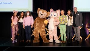 Panel of Disney cast members and creatives pose on stage alongside Duffy and his friend LinaBell at a D23 event celebrating Duffy and Friends, with attendees dressed in themed outfits and the screen behind them displaying event details.