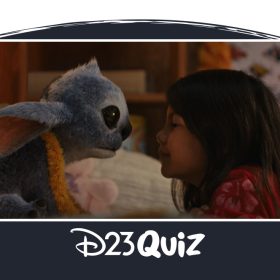 In an image from Disney’s live-action Lilo & Stitch, Stitch (on the left) is sitting on a bed and is facing almost nose-to-nose with Lilo (TK), who’s leaning up against the bed while kneeling on the floor. Stitch is wearing a yellow lei, while Lilo is wearing a pink flowered dress.