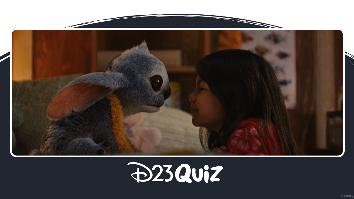 : In an image from Disney’s live-action Lilo & Stitch, Stitch (on the left) is sitting on a bed and is facing almost nose-to-nose with Lilo (TK), who’s leaning up against the bed while kneeling on the floor. Stitch is wearing a yellow lei, while Lilo is wearing a pink flowered dress.