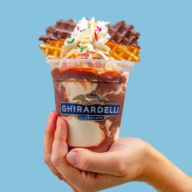 A single hand holds out a Ghirardelli ice cream cup containing vanilla ice cream blended with a generous helping of chocolate fudge syrup. Atop the ice cream are two waffle cones forming Mickey Mouse ears, with some whipped cream topped with sprinkles separating them. The hand holding the sundae is seen in front of a light blue background.
