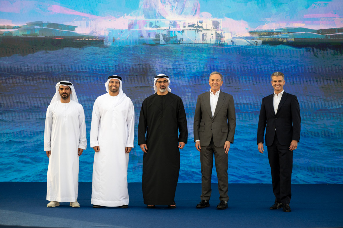 Five executives working on the new Disney theme park resort in Abu Dhabi stand in a line on a stage with a large rendering of Yas Island behind them. The executives include three representatives of Miral, in traditional Arab garb, on the left, and, on the right, Bob Iger and Josh D’Amaro.