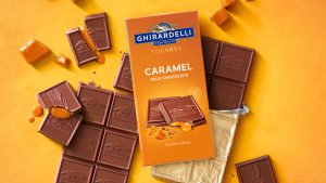Assorted Ghirardelli chocolate squares sit against an orange background. The chocolate squares are stamped with the Ghirardelli logo. A few of the squares are broken open and caramel is trickling from the inside. The front of the package is seen atop the squares; it features the Ghirardelli logo and reads “Squares, Caramel Milk Chocolate, luscious filling.”