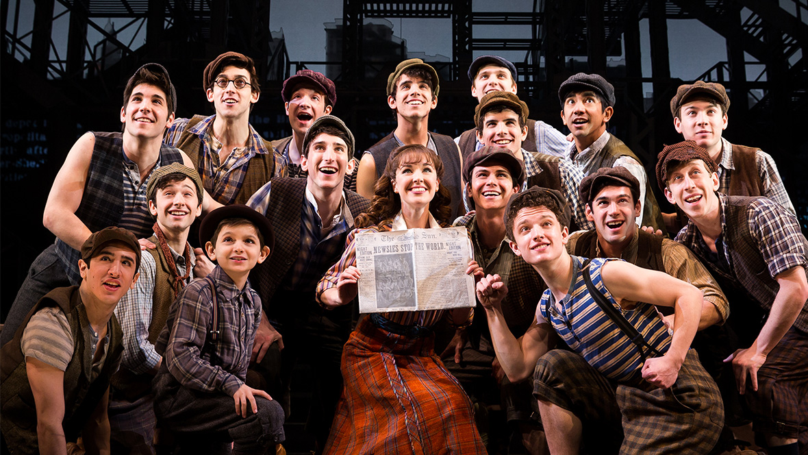 The cast of Newsies: The Broadway Musical huddled around a newspaper talking about their strike and facing the audience while smiling.