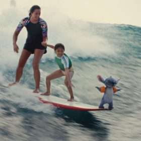 In a scene from Lilo & Stitch (2025), the sisters Nani (Sydney Elizebeth Agudong) and Lilo (Maia Kealoha) ride a surfboard on a tall ocean wave. At the front of the surfboard is Stitch (voiced by Chris Sanders).
