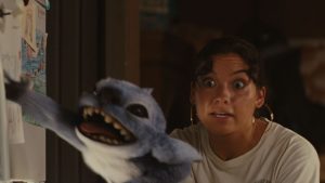In a kinetic scene from Lilo & Stitch (2025), Nani (Sydney Elizebeth Agudong) stands in the kitchen of her house looking and grabbing frantically at Stitch (voiced by Chris Sanders), who is just in front of her, hanging on the open refrigerator door with one paw while reaching for the handle of the upper freezer compartment with the other. Nani’s eyes are wide, and Stitch’s mouth is open, with his tongue sticking out a bit. Behind them is the out-of-focus wall of the kitchen.