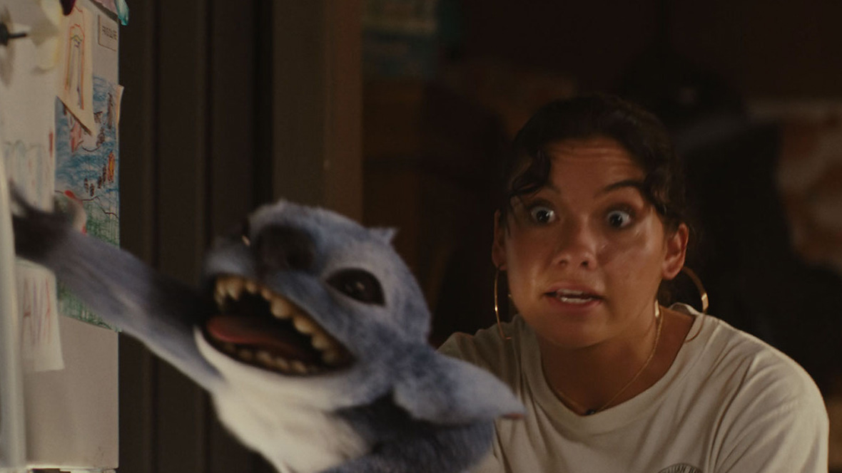 In a kinetic scene from Lilo & Stitch (2025), Nani (Sydney Elizebeth Agudong) stands in the kitchen of her house looking and grabbing frantically at Stitch (voiced by Chris Sanders), who is just in front of her, hanging on the open refrigerator door with one paw while reaching for the handle of the upper freezer compartment with the other. Nani’s eyes are wide, and Stitch’s mouth is open, with his tongue sticking out a bit. Behind them is the out-of-focus wall of the kitchen.