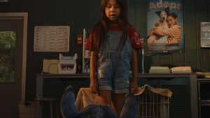 In a scene from Lilo & Stitch (2025), Lilo (Maia Kealoha) stands in front of a counter in the office of a pet adoption agency. She is facing the camera and is seen from just above floor level. In front of her, with his back to the camera, is Stitch (voiced by Chris Sanders), seen only from the neck up. Lilo is wearing denim overall shorts and a red shirt. Behind her on the counter are stacks of paper and other common office items. A door with a window to the outside can be seen on the left, and on the right is a poster on the wall of a girl hugging a dog and the words &ldquo;Adopt &ndash; Save a Life.&rdquo;