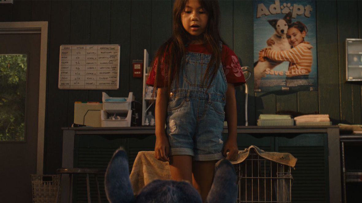 In a scene from Lilo & Stitch (2025), Lilo (Maia Kealoha) stands in front of a counter in the office of a pet adoption agency. She is facing the camera and is seen from just above floor level. In front of her, with his back to the camera, is Stitch (voiced by Chris Sanders), seen only from the neck up. Lilo is wearing denim overall shorts and a red shirt. Behind her on the counter are stacks of paper and other common office items. A door with a window to the outside can be seen on the left, and on the right is a poster on the wall of a girl hugging a dog and the words “Adopt – Save a Life.”