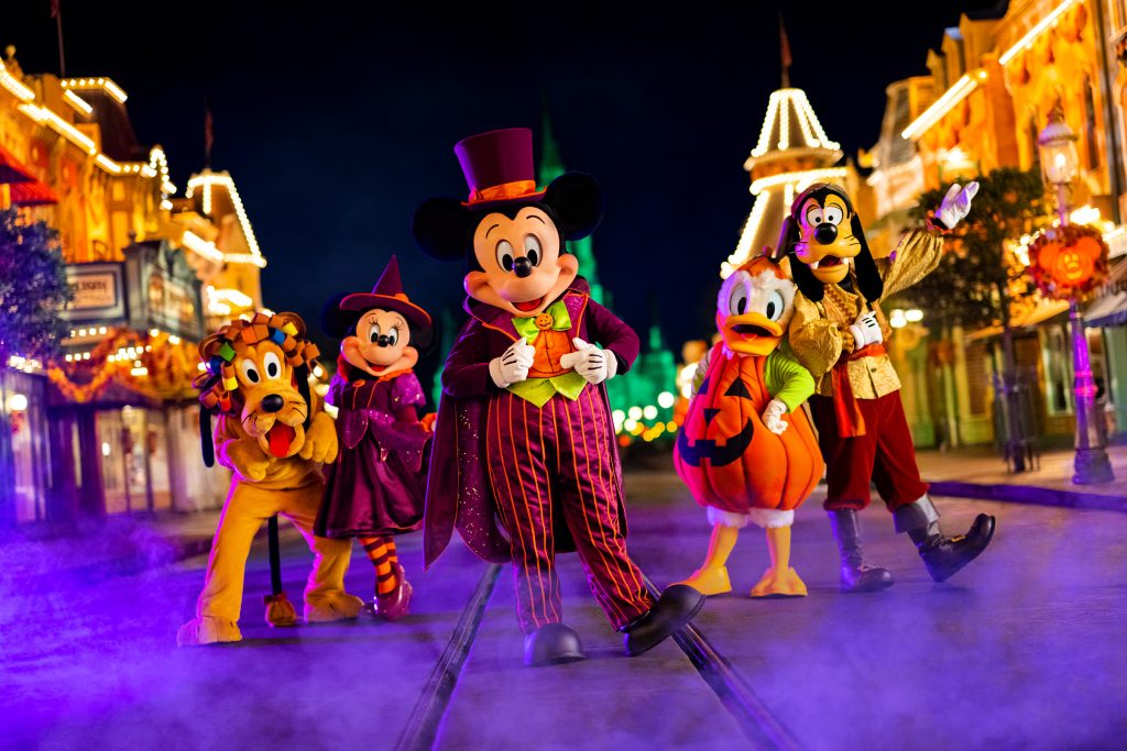 Mickey Mouse and friends dressed in Halloween costumes pose on a foggy, festively lit Main Street at night. Mickey wears a purple top hat and striped suit, Minnie is a cheerful witch, Donald is dressed as a pumpkin, Goofy wears a skeleton-themed suit, and Pluto has a lion’s mane. Orange lights and Halloween decorations line the buildings in the background, capturing the spooky fun spirit of Mickey’s Not-So-Scary Halloween Party.