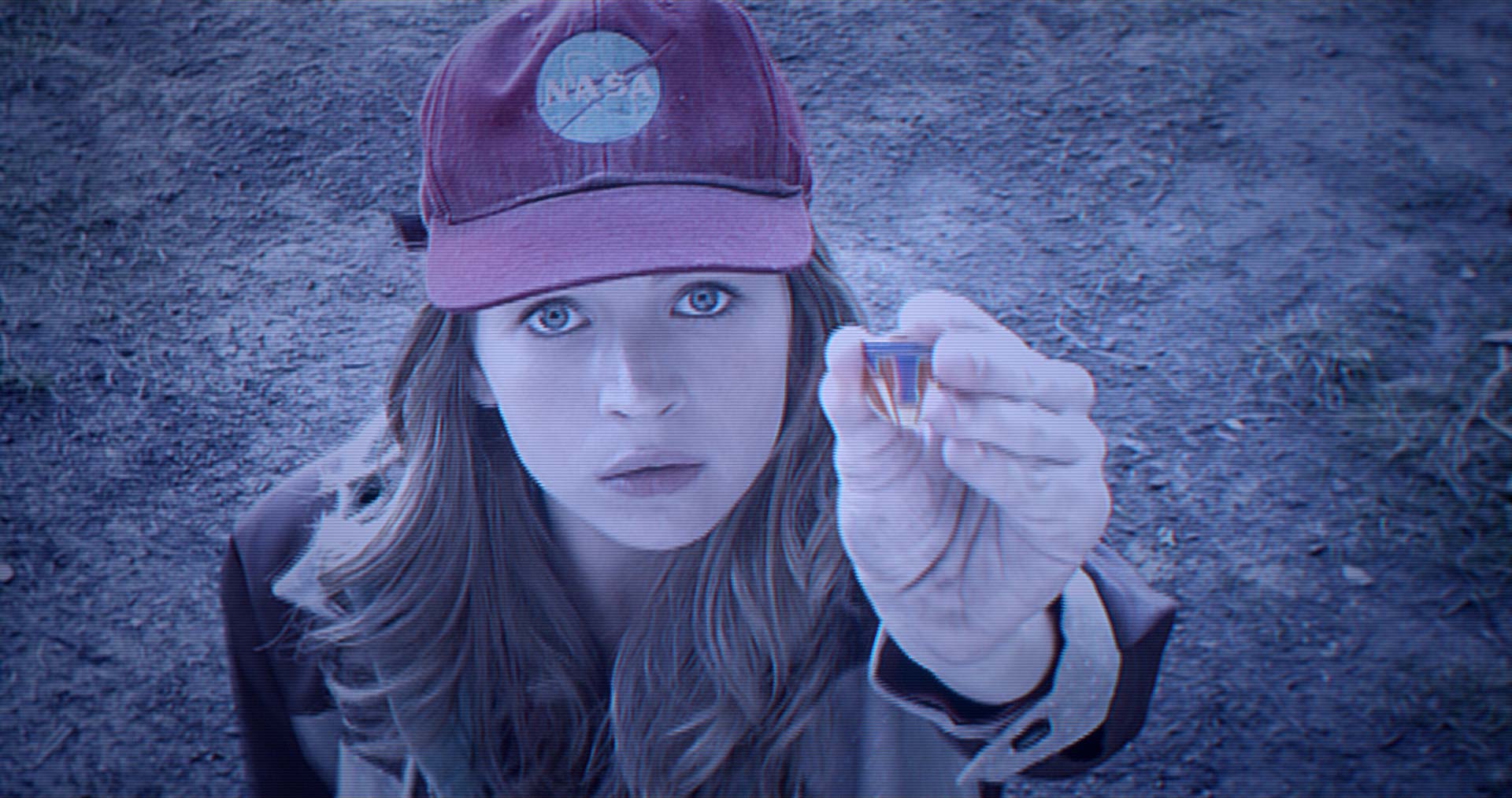A young woman wearing a red NASA cap gazes upward with wide eyes, holding a glowing pin between her fingers. The image appears to be seen through a screen, with scan lines adding a retro, surveillance-style effect.