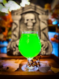 The Poison Green Apple Seltzer from Unsung Brewing. Pictured is a glass with bright, green-colored liquid, highlighted in front of a skull backdrop.