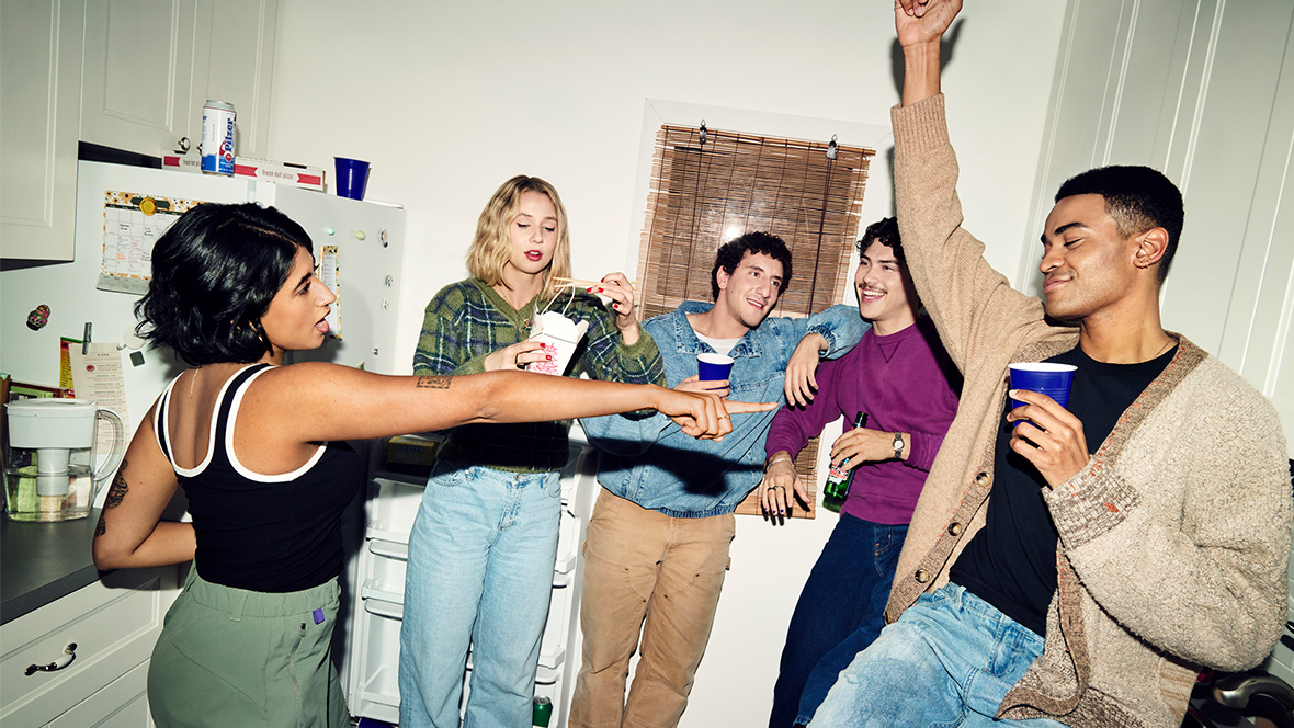 The main ensemble cast of FX’s Adults standing in a white kitchen, drinking out of beer bottles and plastic cups. One member is eating noodles out of a white to-go box.